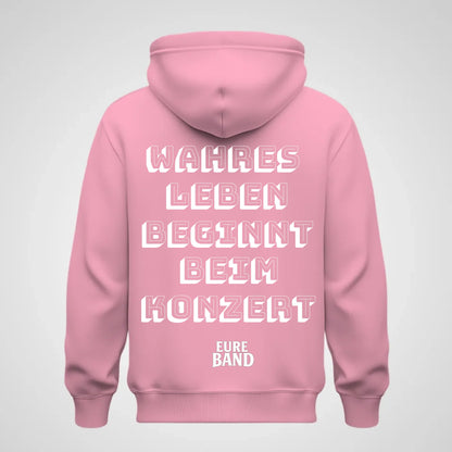 Rosa Hoodie mit großem weißen Schriftzug auf der Rückseite: ‚Wahres Leben beginnt beim Konzert‘. Darunter steht der Schriftzug ‚Eure Band‘. Der Bandname kann individuell angepasst werden.